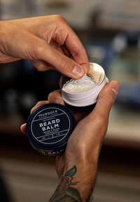 Skæg balm i en mørkeblå dåse med hvid tekst, åben for at afsløre et cremet hvidt produkt under et sølvbeskyttelseslåg, holdt i hænderne.