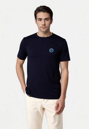 Marineblaue kurzärmelige T-Shirt aus glattem Stoff. Mit einem kleinen blauen Logo auf der linken Brust. Trägt helle Hose.