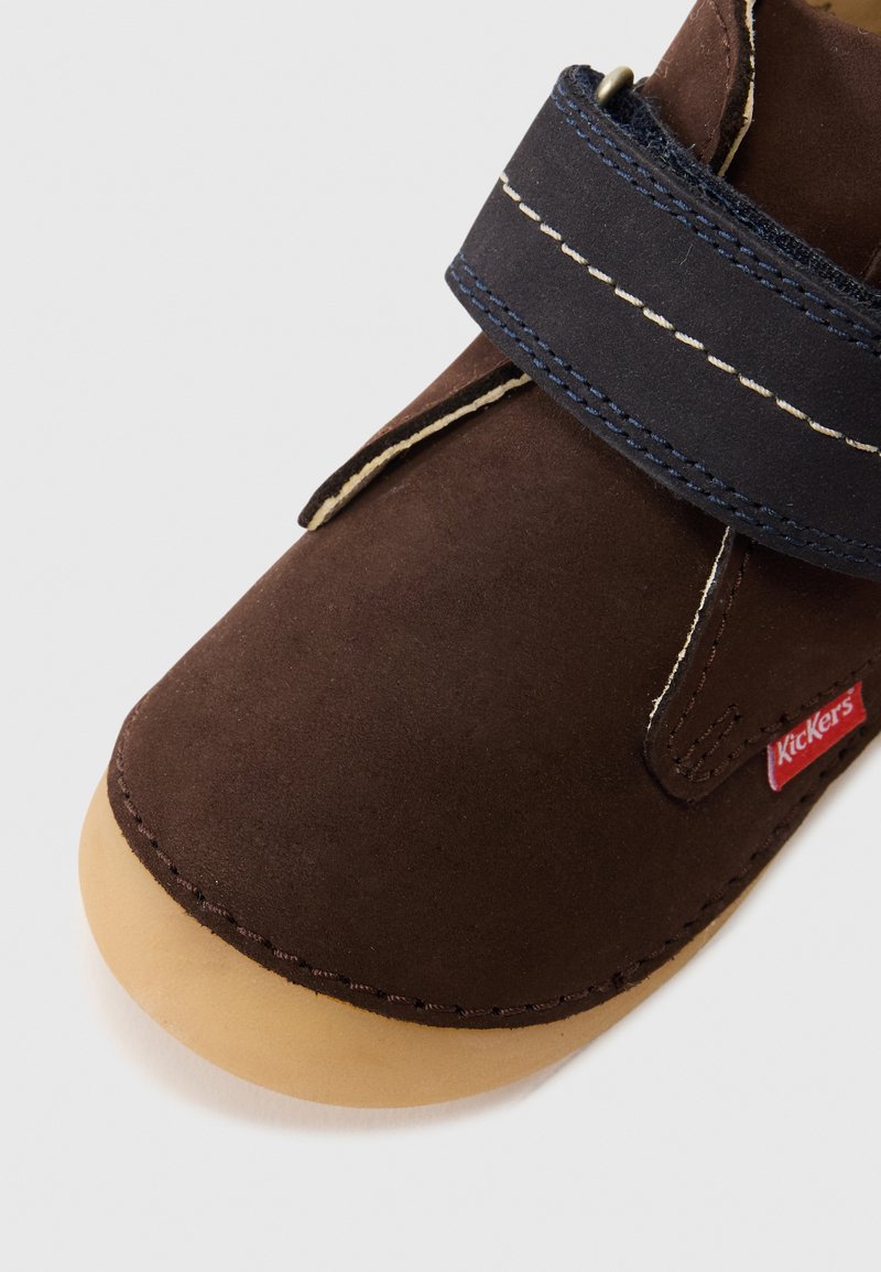 Scarpa in suede marrone con punta rotonda, suola in gomma e una larga strap in velcro blu navy. Presenta cuciture a contrasto color crema e un'etichetta logo rossa.