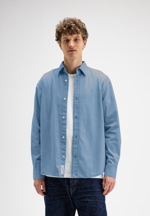 Junger Mann mit lockigem Haar, der ein hellblaues Hemd über einem weißen T-Shirt und dunklen Jeans trägt, steht vor einem schlichten hellen Hintergrund.
