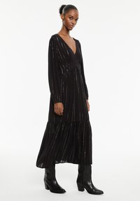 Abito maxi nero con scollo a V profondo, maniche lunghe e orlo a strati. Presenta strisce metalliche verticali e un tessuto fluido e testurizzato.