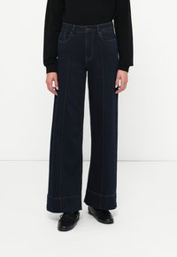 Jeans de pierna ancha de color azul oscuro con una cintura alta, que presentan costuras en contraste y bolsillos frontales, combinados con zapatos negros.