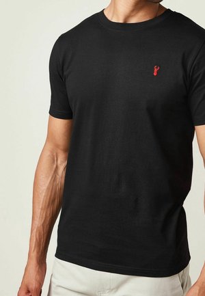 Homme portant un t-shirt noir ajusté à manches courtes avec un petit logo tête de cerf rouge sur la poitrine gauche, associé à un pantalon de couleur claire.