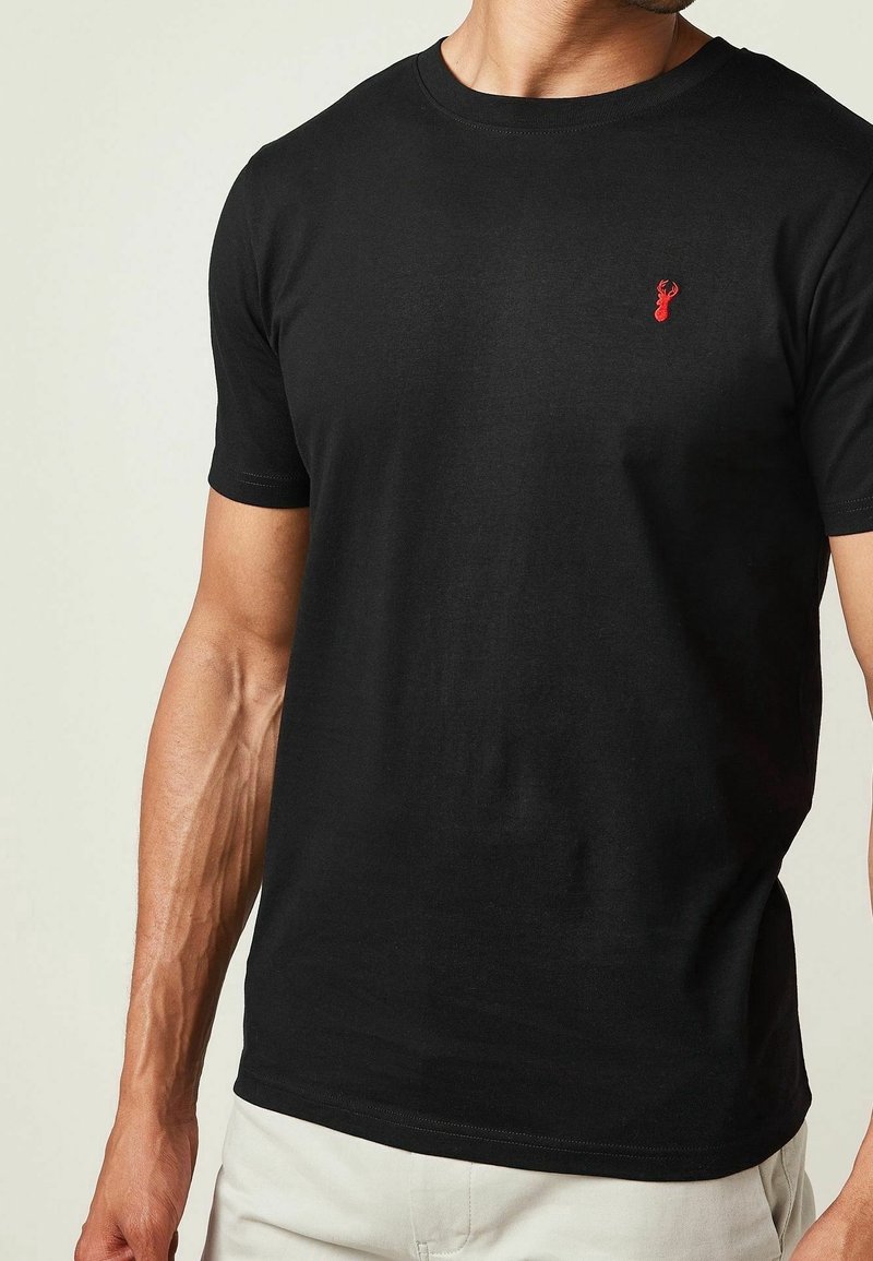 Homme portant un t-shirt noir ajusté à manches courtes avec un petit logo tête de cerf rouge sur la poitrine gauche, associé à un pantalon de couleur claire.