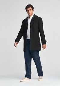 Svart yllerock med huva, en knappknäppning, sidofickor, kombinerad med ljusa denimjeans och krämfärgade sneakers. Enkel, strukturerad design.