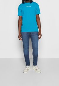 Camiseta turquesa brillante con un pequeño logo blanco y rojo en el pecho, combinada con jeans ajustados azules y zapatillas blancas con detalles beige.
