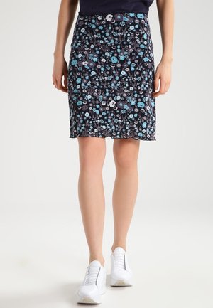 Vrouw draagt een zwarte, bloemenknielange rok met blauwe en witte bloemen, gecombineerd met witte sneakers, staand tegen een lichte achtergrond.