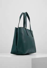 Tote bag nahkaa tummanvihreää tekstuuria, jossa on kaksi olkahihnaa, seisoo valkoisella jalustalla harmaata taustaa vastaan.