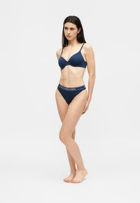 Femme debout pieds nus, portant un soutien-gorge bleu marine et une culotte assortie avec une ceinture "Polo Ralph Lauren", sur un fond clair uni.