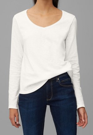 Vrouw die een wit, lange mouwen, V-hals shirt draagt, lichtjes ingestopt in een donkerblauwe spijkerbroek, tegen een grijze achtergrond.