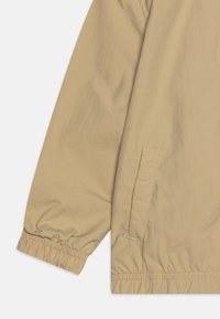 Ljusbeige jacka med en slät yta, elastiska ärmslut och en sidoficka. Har enkel sömnad och en avslappnad passform.