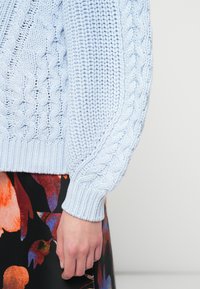 Maglione azzurro chiaro a maglia lavorata a trecce con motivi texturizzati e polsini a coste, abbinato a una gonna floreale multicolore.