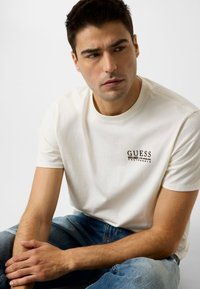 T-shirt en coton blanc à col rond, avec un petit logo "GUESS" marron sur la poitrine gauche. Le modèle porte un jean denim bleu.