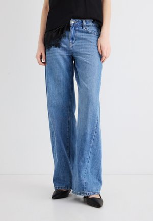 Femeie purtând blugi largi de culoare albastru deschis, top negru fără mâneci și pantofi negri cu vârf ascuțit, stând pe un fundal alb.