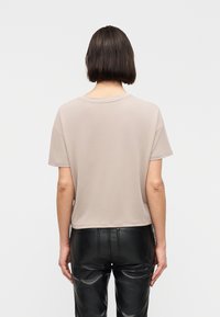 Άτομο με κοντά σκουρόχρωμα μαλλιά, που φοράει μπεζ κοντομάνικο πουκάμισο και μαύρο δερμάτινο παντελόνι, στέκεται πλάτη σε απλό φόντο.