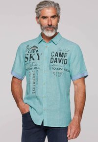 Turquoise kortemouwen shirt met knoopsluiting aan de voorkant, met bedrukte tekst en een lichtblauw accent op de mouwen. Gewaxt stof.