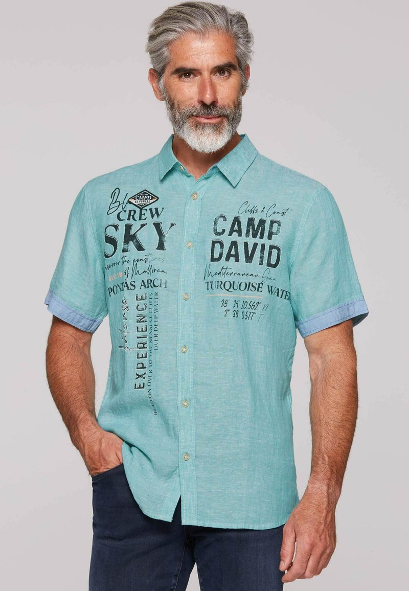 Turquoise kortemouwen shirt met knoopsluiting aan de voorkant, met bedrukte tekst en een lichtblauw accent op de mouwen. Gewaxt stof.