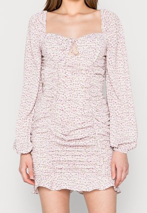 Femme portant une mini-robe rose clair froncée avec de longues manches bouffantes et un décolleté carré avec un détail de nouage devant.