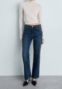 Suéter ajustado de manga larga en beige claro con cuello alto, combinado con jeans azul de pierna recta de cintura alta. Tacones negros de punta afilada complementan el atuendo.