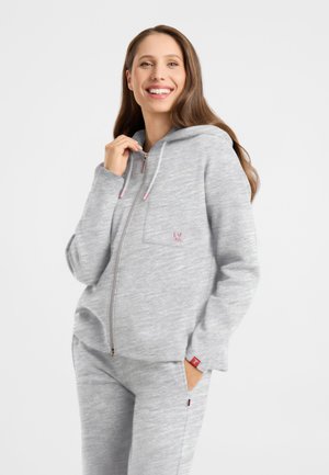 Smiling woman wearing light grey zip-up hoodie and matching sweatpants, standing with one hand in pocket and the other near the zipper.