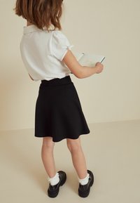 Schwarzer, ausgestellter Rock mit einem taillierten Bund, kombiniert mit einem kurzärmeligen weißen Poloshirt. Das Kind hält ein Notizbuch und schaut weg.
