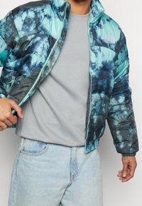 Hombre con una chaqueta de plumas de color azul claro y efecto tie-dye sobre una camiseta gris y jeans azules claros, ajustando la cremallera de la chaqueta con la mano izquierda.