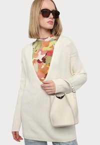 Kreemi neulecardigan syvällä V-kaula-aukolla, joka on puettu monivärisen kamouflagepaidankin päälle. Mallilla on kädessään pieni, vaaleanvärinen käsilaukku.