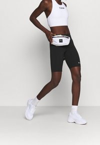 Czarne spodenki kolarskie w połączeniu z białym sportowym biustonoszem i białymi trampkami. Mała biała nerka z logo znajduje się w talii.