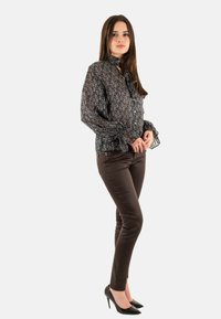 Blusa transparente con estampado negro y azul, con cuello acampanado y mangas con puños, combinada con pantalones marrones ajustados y tacones altos negros.