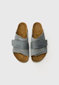 Sandales en daim gris avec une large sangle et une boucle ajustable. Semelle intérieure texturée marron et semelle en caoutchouc. Logo de la marque embossé sur la sangle.