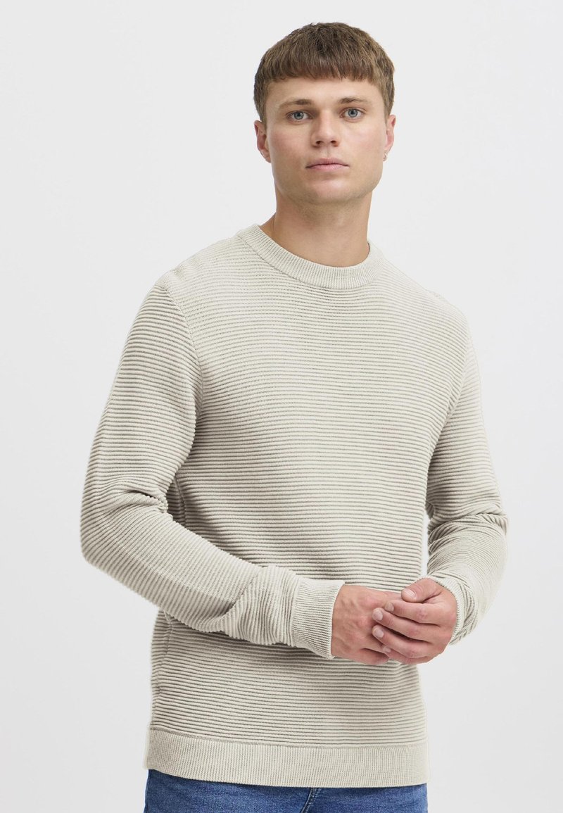 Jaunas vyras trumpais rudu plaukais, dėvintis šviesiai smėlio spalvos ribinę megztinį ir mėlynas džinsas, stovintis priešais paprastą baltą foną.