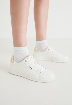 Baskets blanches avec une finition lisse, présentant un accent doré au niveau du talon, un laçage à l'avant et un design minimaliste. Associées à des chaussettes blanches.