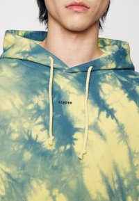 Jeune personne portant un sweat à capuche tie-dye bleu et jaune avec le texte "SANDRO" et des cordons blancs, cadré au niveau du cou et des épaules.