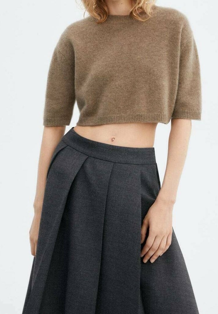 Femme portant un pull court marron et une jupe plissée foncée taille haute, debout devant un fond clair uni.
