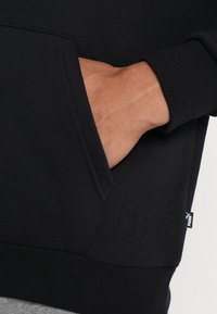 Sweatshirt noir avec poche kangourou, poignets et ourlet côtelés. Présente une petite étiquette blanche Puma sur le côté. Texture douce.