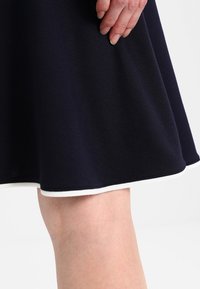 Gonna blu navy con orlo bianco, realizzata in tessuto morbido; presenta una consistenza liscia e un design svasato; mano visibile appoggiata sul tessuto.