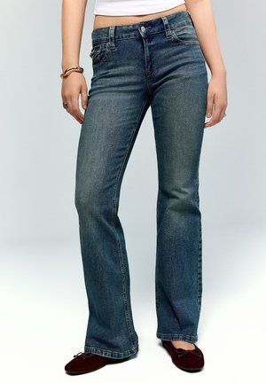 Žena v tmavomodrých bootcut džínsach, tmavohnedých baletných topánkach, bielom crop tope a náramkoch na ľavom zápästí, stojaca na jednofarebnom pozadí.