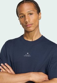 Námornícky modré bavlnené tričko s okrúhlym výstrihom a krátkymi rukávmi. Na hrudi je potlačené biele logo "Adidas Originals". Jednoduchý dizajn.