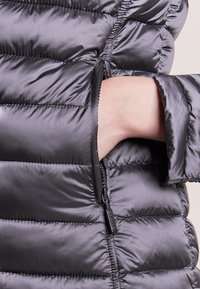 Schwarze Puffjacke mit gesteppten Abschnitten, glatter Textur und einer seitlichen Reißverschlusstasche für praktische Aufbewahrung.
