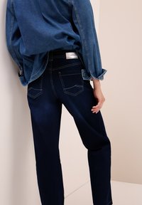 Persona vistiendo jeans de denim azul oscuro y una camisa de denim a juego con las mangas arremangadas, de pie frente a una pared beige.