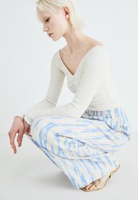 Light cream ribbed long-sleeve top with a V-neck, paired with a flowing skirt featuring a blue and cream abstract pattern. Gold flat sandals.