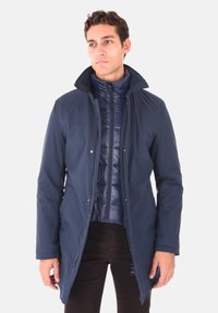 Cappotto lungo blu navy con colletto alto, chiusure a bottone e uno strato interno trapuntato. Dotato di tasche laterali e una texture liscia e resistente.
