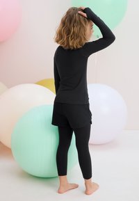 Vaiko dėvimas juodas ilgomis rankovėmis megztinis ir šortai su juodomis pėdkelnėmis, pasižymintis glotniu audiniu, fone pasteliniai balionai.