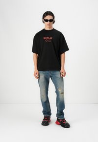 Maglietta nera oversize con logo rosso "REPLAY", jeans chiari strappati e sneaker chunky nere e rosse. Modello con occhiali da sole neri.