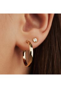 Goldene Creolen mit polierter Oberfläche, kombiniert mit einem kleinen Stecker mit klarem Stein, auf dem Ohrläppchen gegen die Haut präsentiert.