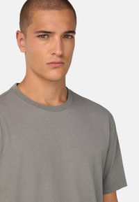 T-shirt gris à col rond fabriqué en coton doux. Manches courtes, coupe standard avec une texture subtile. Visage du modèle partiellement visible avec une expression neutre.