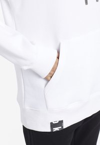 Mano nella tasca di una felpa bianca con polsini a costine e un'etichetta nera visibile, abbinata a pantaloni neri su uno sfondo chiaro.