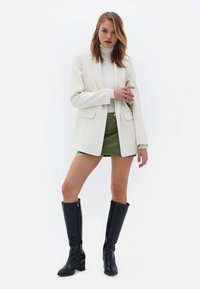 Blazer bianco, dolcevita a coste, mini gonna verde e stivali neri fino al ginocchio. Il blazer presenta due tasche frontali e una vestibilità aderente.