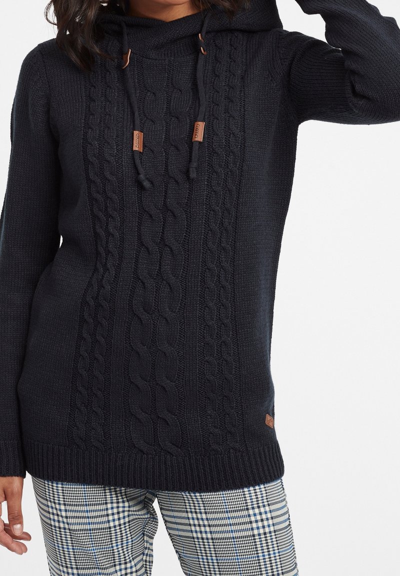 Pullover in lana di un blu scuro, lavorato a maglia con motivo a treccia, cappuccio e lacci. Presenta dettagli in pelle e polsini e orlo a coste. Abbinato a pantaloni a quadri.