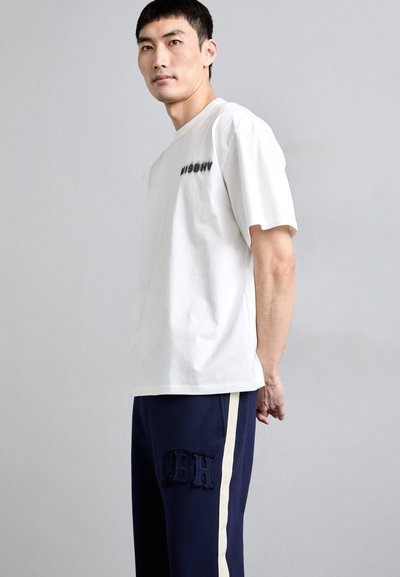 T-shirt en coton blanc avec "MISBHV" imprimé en noir sur le devant. Pantalons de survêtement bleu marine avec des rayures crème sur les côtés et broderie "MBH" sur la cuisse.